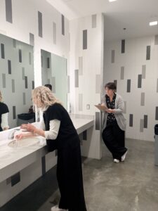 Researcher holding a clipboard observes subject washing her hands in the bathroom. Another researcher is mostly hidden from view using binoculars to observe the subject.