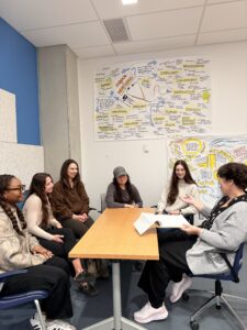 A group of six people sit around a table in a classroom. A researcher leads the discussion, holding a sheet of paper while the others listen and collaborate.
